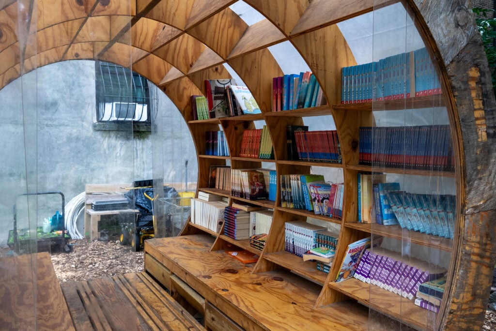 A cozy wooden library space features curved shelves filled with colorful books. The transparent walls allow natural light, enhancing the inviting atmosphere. A wooden bench runs along the front, providing a place to sit and read.