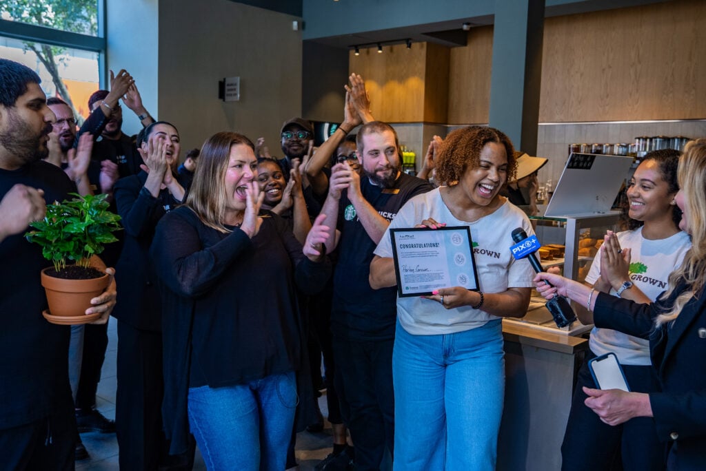A group of people is celebrating in a café, clapping and smiling. One woman holds a certificate labeled "CONGRATULATIONS!" while being interviewed. The atmosphere is joyful and festive.
