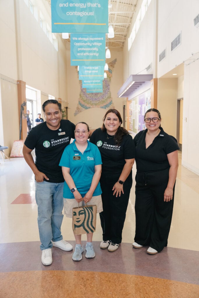 Four individuals pose together in a brightly lit hallway. Two wear black shirts with the Starbucks Foundation logo, while one wears a teal shirt and the other is in black. The person in teal holds a small Starbucks bag. Colorful banners hang from the ceiling, and a decorative wall is visible in the background.