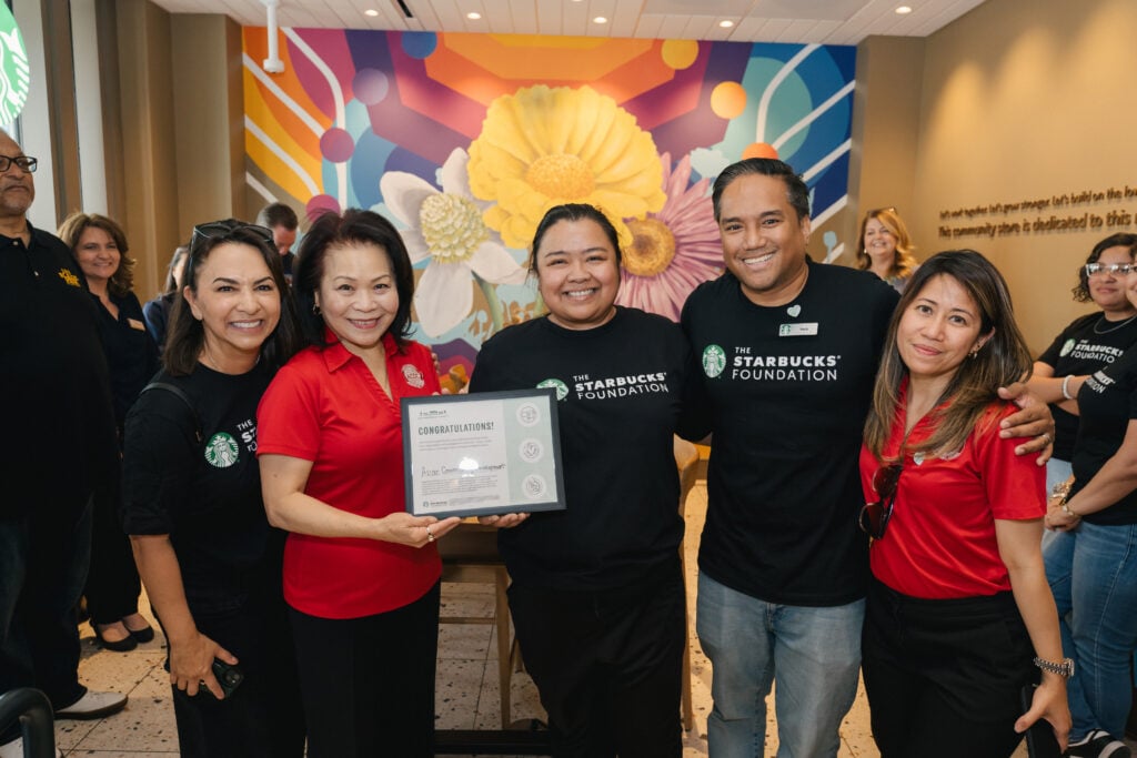A group of five people poses together, smiling and holding a certificate that reads "CONGRATULATIONS!" The background features a colorful mural with flowers.