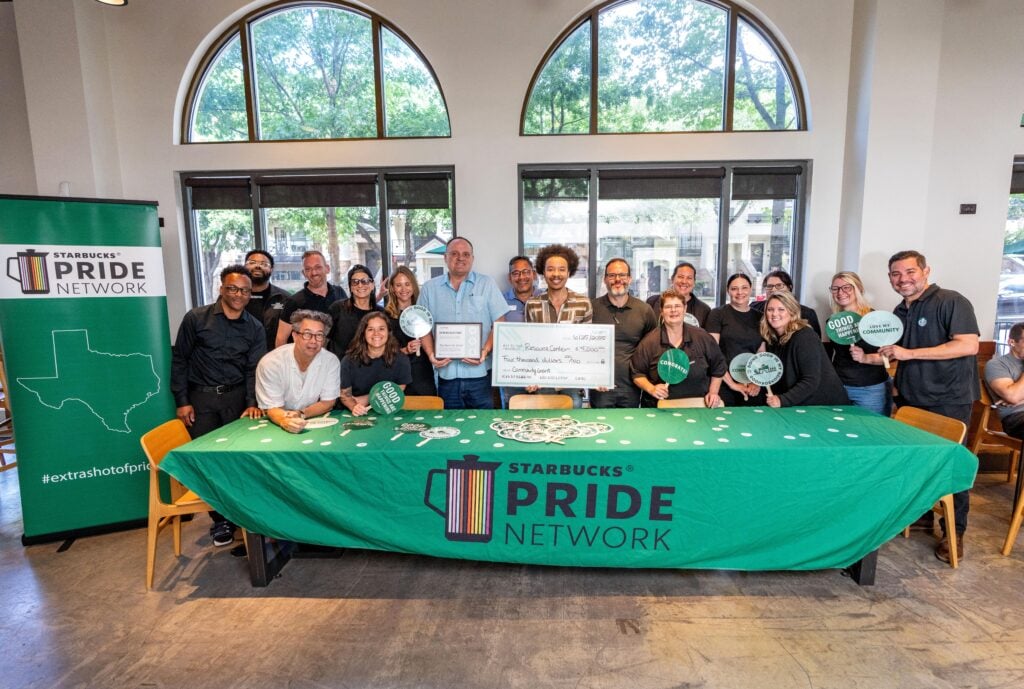 A group of people stands around a table covered with a green Starbucks Pride Network tablecloth. They are holding a large check for $4,000 made out to a resource center. The atmosphere is celebratory, with various signs and smiles among the participants.