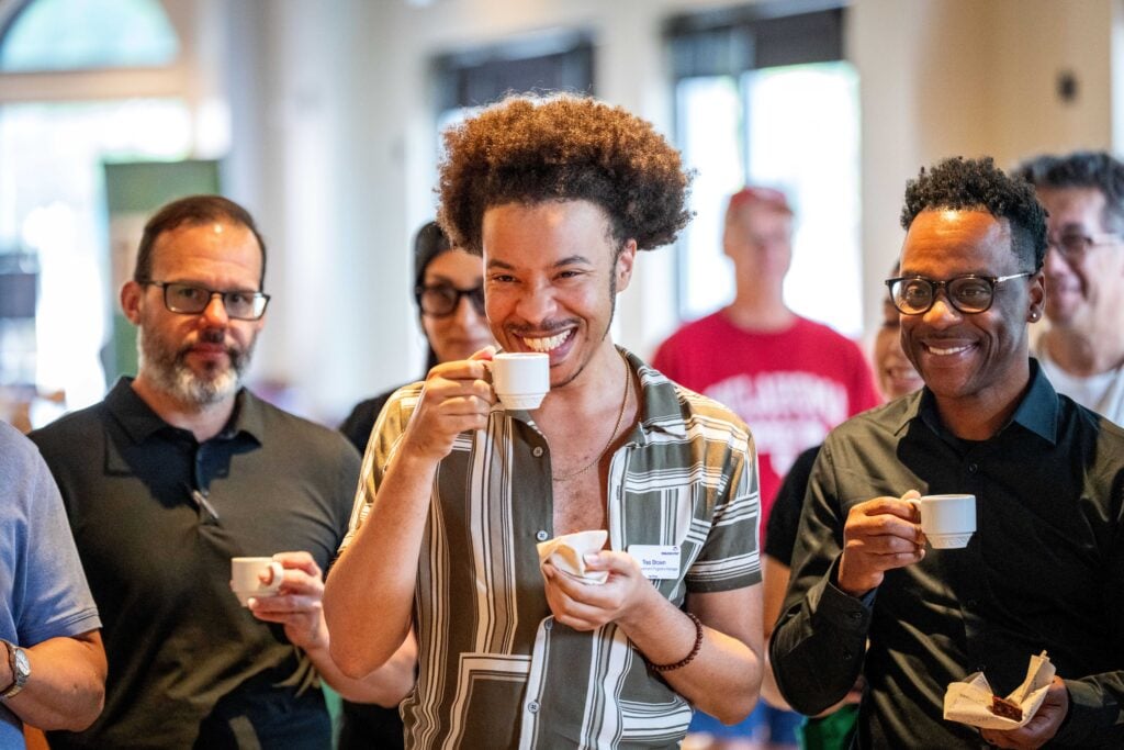 A group of people enjoying drinks, with one man in the foreground smiling and holding a small cup. He has curly hair and is wearing a striped shirt. Others in the background are also holding cups.