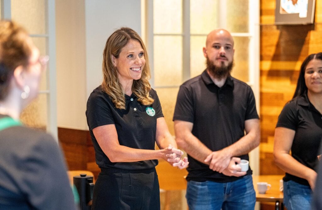 A woman with long, wavy hair smiles while speaking, wearing a black polo shirt with a logo. A man with a beard stands beside her, both in a casual setting.