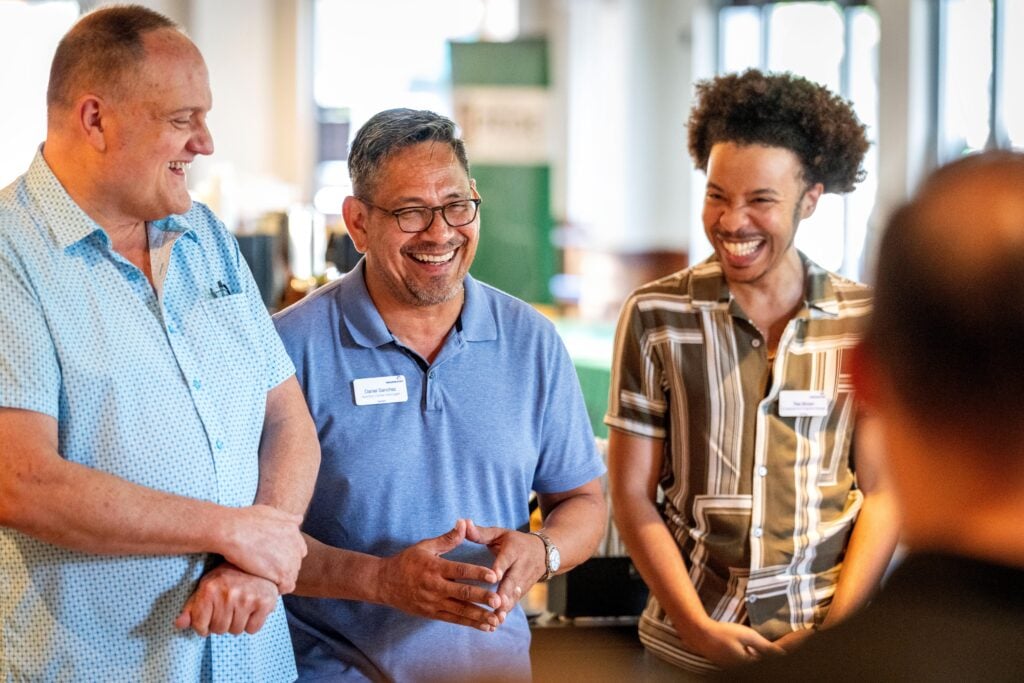 Three men are smiling and engaged in conversation. One wears a light blue polo shirt, another has a patterned shirt, and the third is in a striped shirt. They appear to be enjoying a friendly moment in a bright, casual setting.