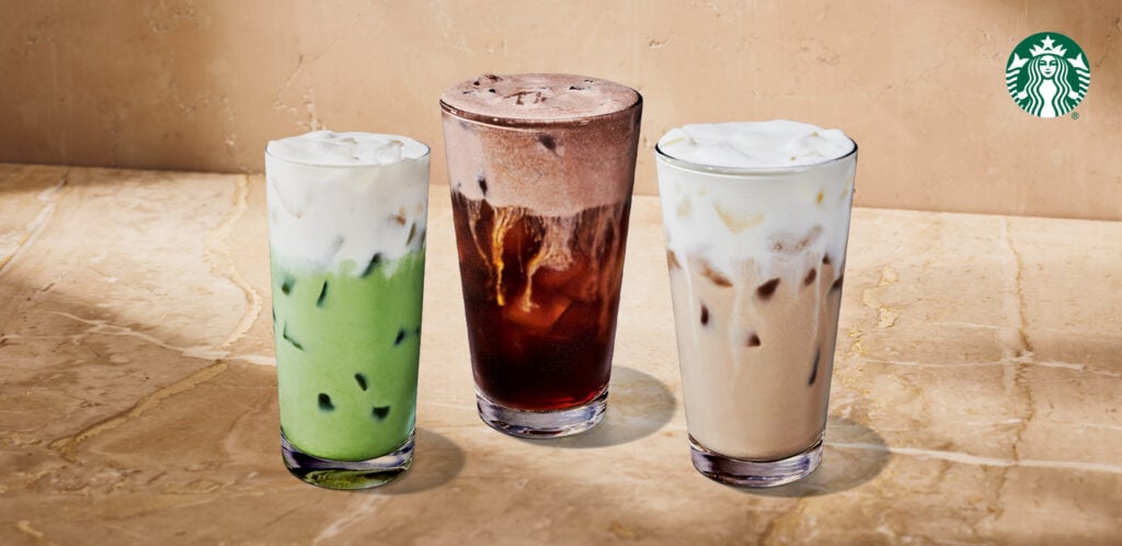 Three iced beverages are displayed on a marble surface. From left to right: a green drink with a creamy top, a dark iced drink with a frothy layer, and a light brown drink with a similar frothy top.
