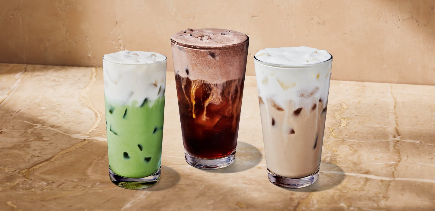 Three tall glasses of iced beverages are displayed on a marble surface. The left glass contains a green drink topped with foam, the center glass has a dark iced drink, and the right glass features a light-colored iced beverage with foam.
