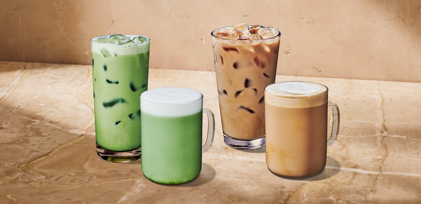 Four iced beverages are displayed on a marble surface. From left to right: a tall glass of green iced drink, a frosted mug with a light green drink topped with foam, a tall glass of iced coffee, and a mug of creamy iced coffee.