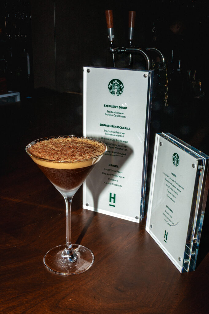 A martini glass filled with a dark espresso cocktail topped with a frothy layer and sprinkled with cocoa. Nearby, a menu displays various drinks and cocktails.