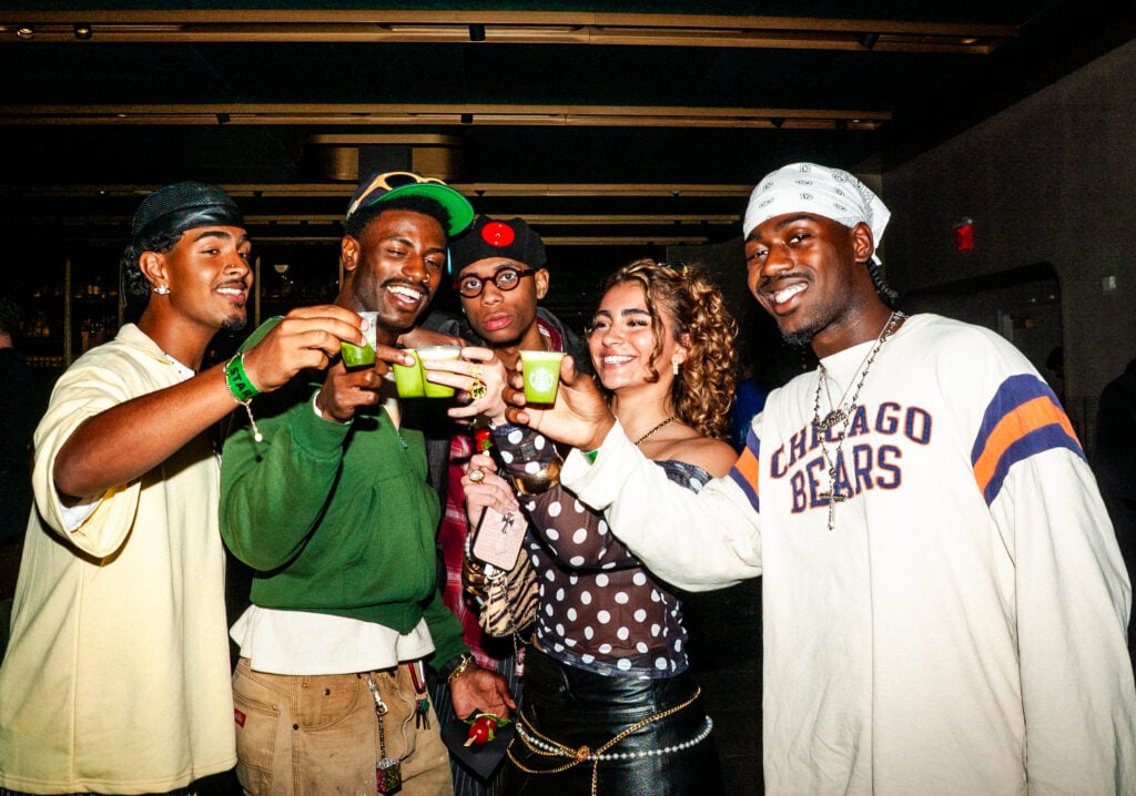 A group of five friends is gathered, smiling and raising colorful shot glasses. They are dressed in casual, trendy outfits, with accessories like hats and jewelry. The setting appears lively, suggesting a celebration or party atmosphere.