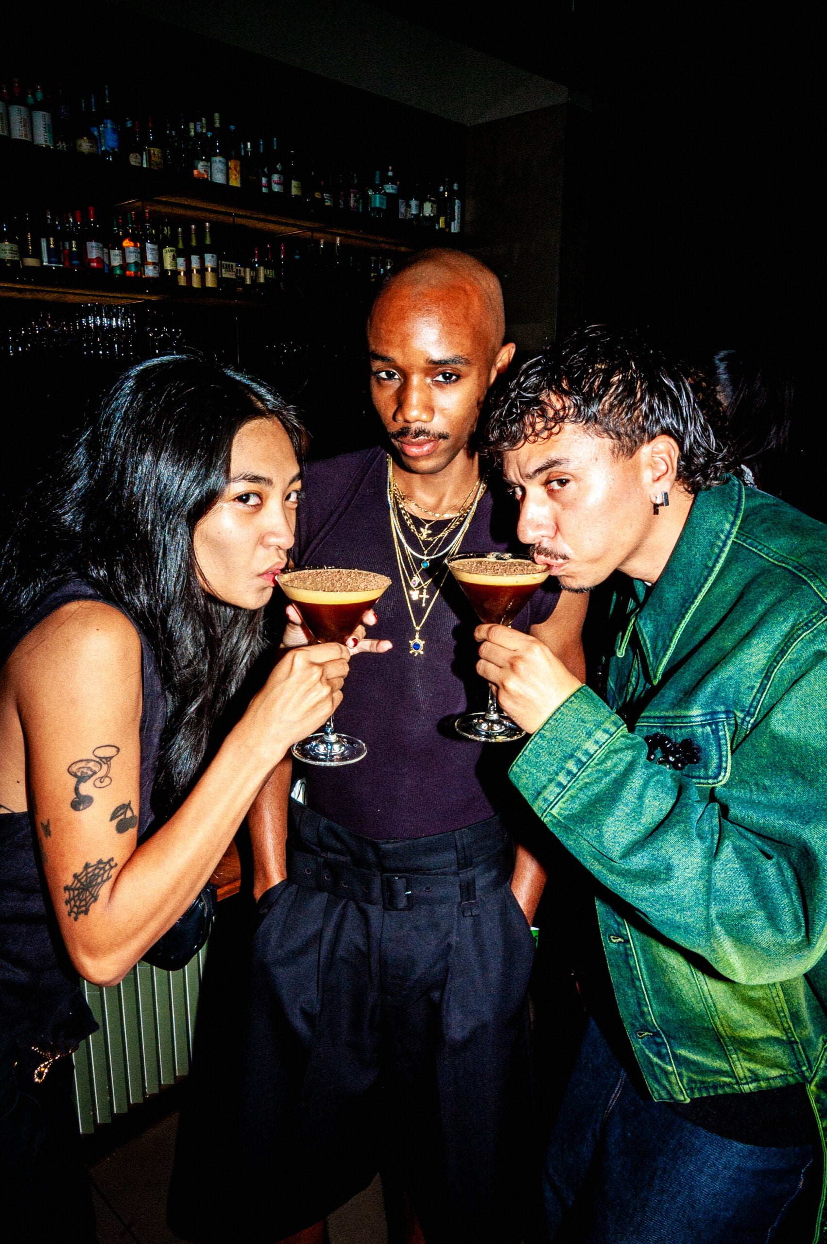 Three individuals pose together, each holding a cocktail in a dimly lit bar. The person on the left has long black hair and a tattoo on their arm, while the middle person has a bald head and wears multiple gold necklaces. The person on the right sports curly hair and a green denim jacket. The background features shelves lined with various bottles.
