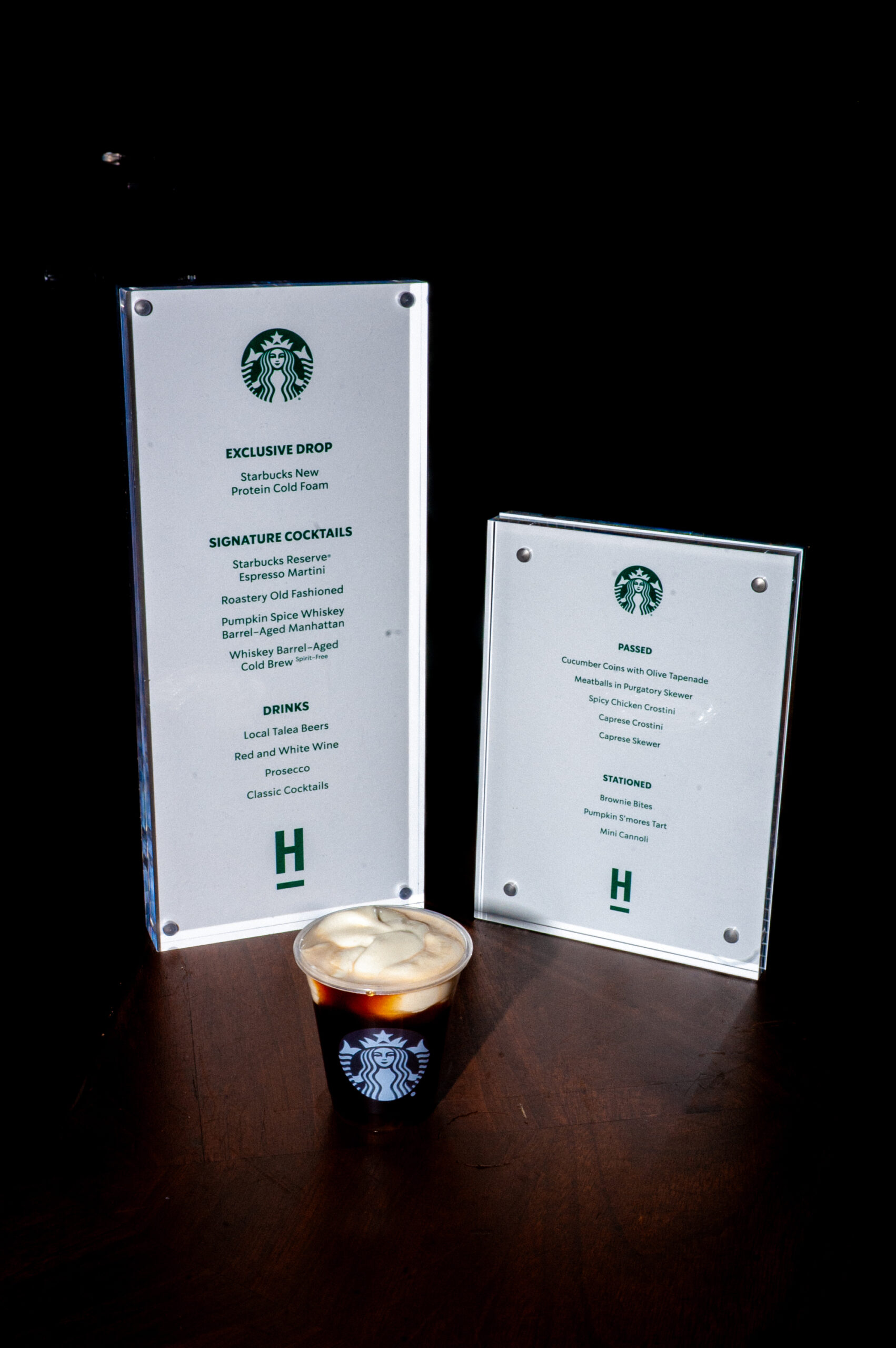Two clear menu stands display food and drink offerings, including Starbucks cocktails and appetizers, alongside a cup of cold brew topped with foam.