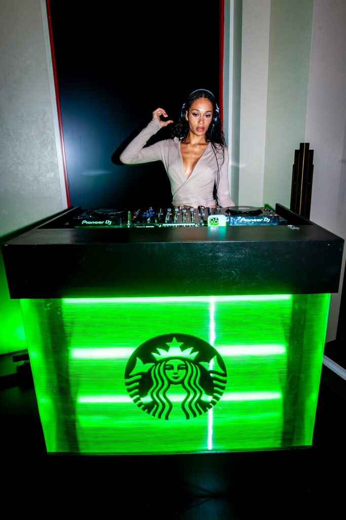 A DJ stands behind a glowing green DJ booth featuring the Starbucks logo. She wears headphones and has long, wavy hair.