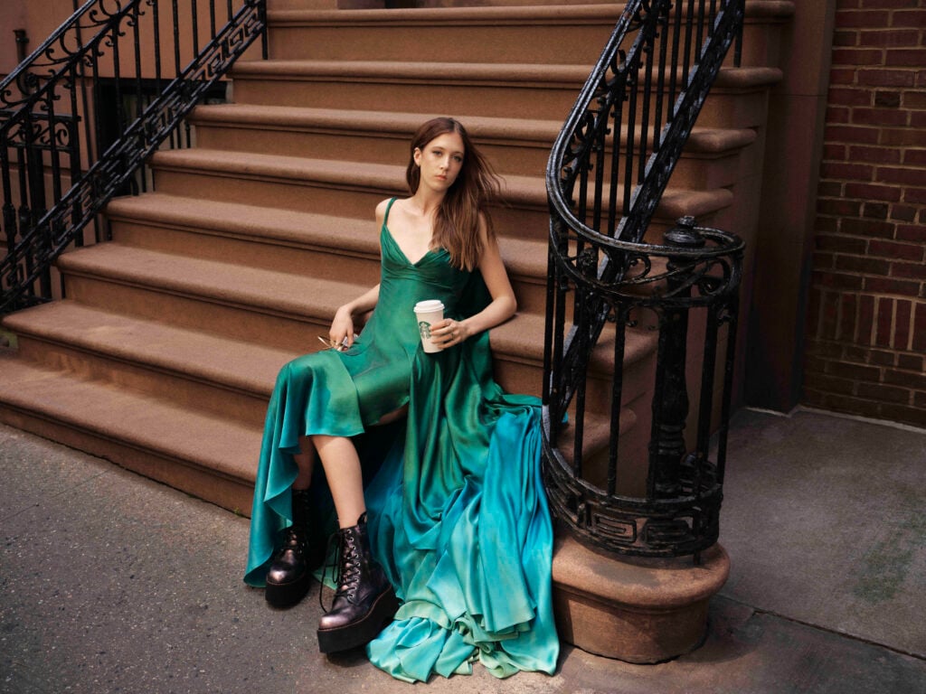 A model sits on brown stone steps, wearing a flowing green dress with an ombre effect. She holds a coffee cup and wears chunky black boots. A decorative iron railing is visible beside her.