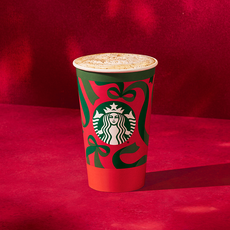 A festive Starbucks cup featuring a green and red design with bows, topped with a frothy beverage. The background is a rich red.