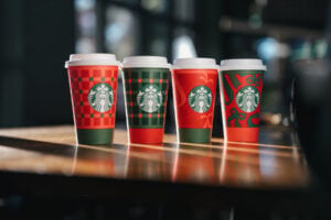 Four festive Starbucks cups are displayed on a wooden table, featuring various holiday-themed designs in red and green.