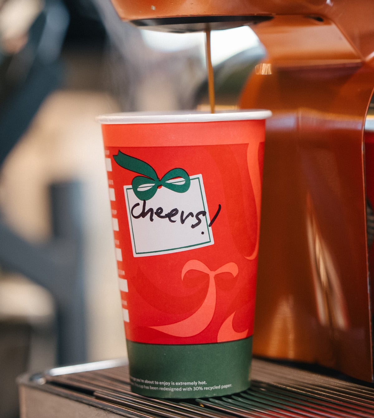 A festive red coffee cup with a green base features a decorative bow and the word "cheers!" written on it. Coffee is being poured into the cup.