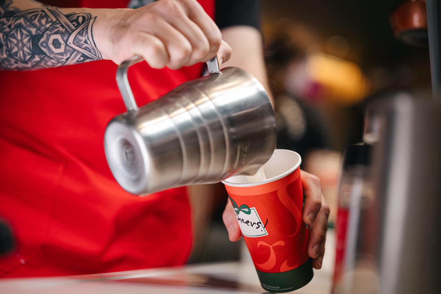 A barista in a red apron pours steamed milk from a metal pitcher into a festive red cup, creating a coffee drink.