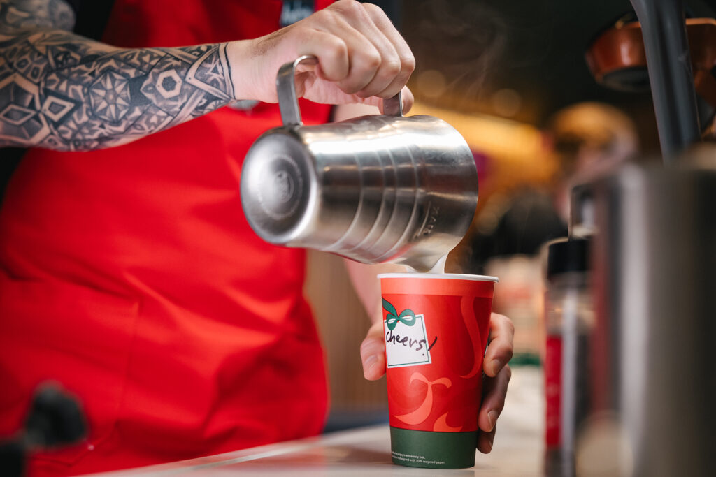 A barista in a red apron pours steamed milk from a metal pitcher into a festive red cup labeled "Cheers!" with steam rising.