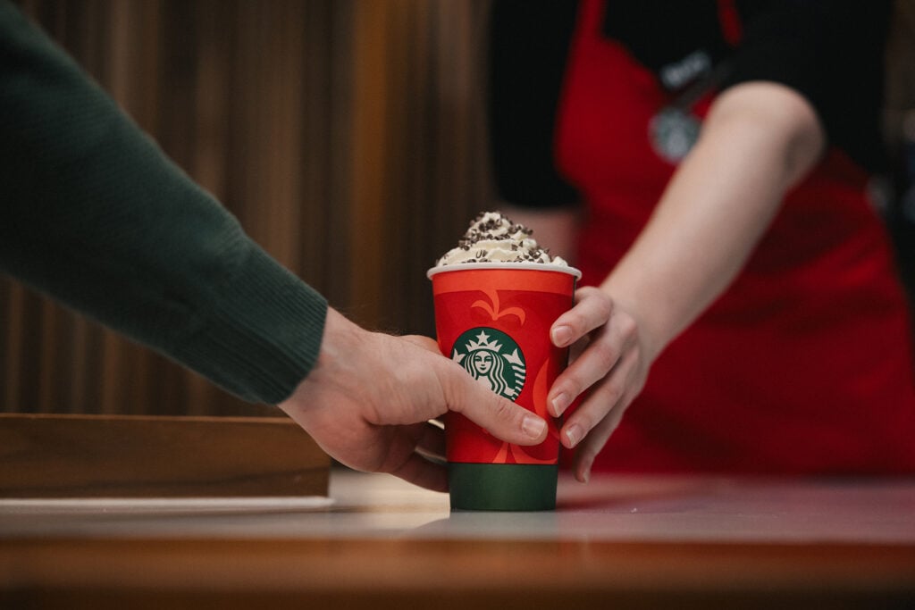 A hand reaches for a red Starbucks cup topped with whipped cream and chocolate shavings, while another hand offers it.