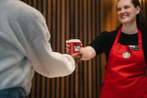 A barista in a red apron hands a festive red coffee cup to a customer. The background features wooden paneling, creating a warm atmosphere.