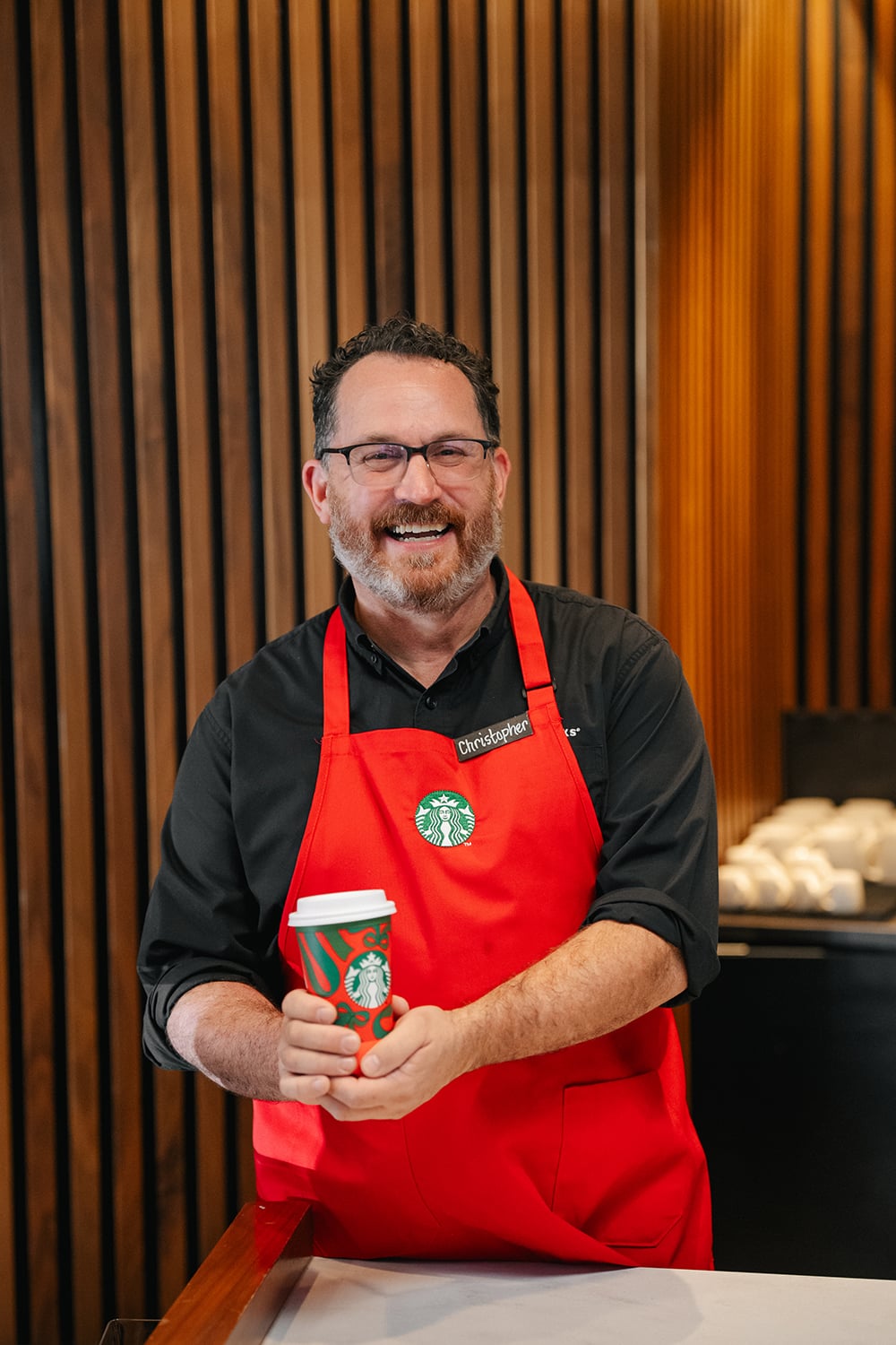 A smiling barista in a red apron holds a festive Starbucks cup in front of a wooden backdrop, showcasing a warm atmosphere.