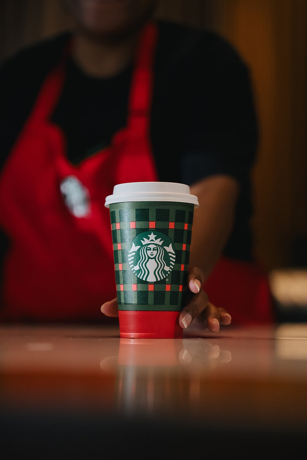 A Starbucks cup with a festive green and red plaid design is being held by a barista wearing a red apron.