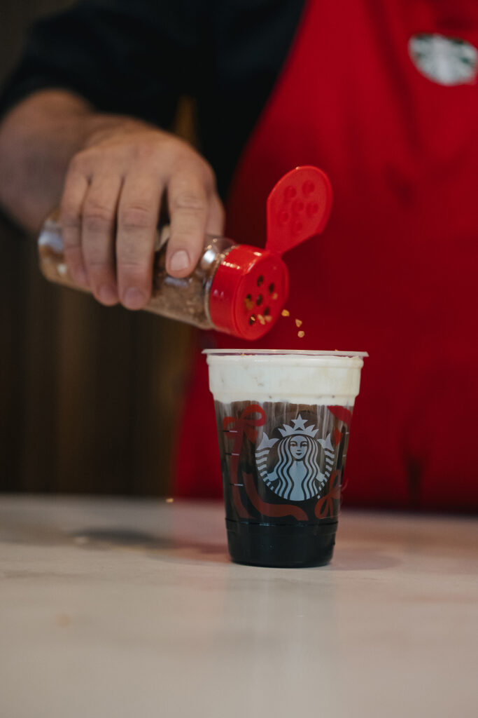 A hand is sprinkling a topping from a shaker onto a Starbucks cup filled with dark liquid and topped with foam. The background is blurred.
