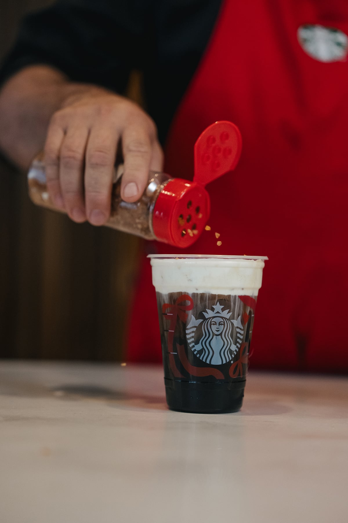A hand is sprinkling a topping from a shaker onto a Starbucks cup filled with dark liquid and topped with foam. The background is blurred.