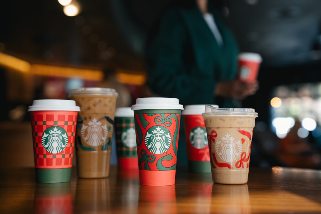 A variety of Starbucks coffee cups are displayed on a wooden table, featuring festive designs in red and green. Some cups are hot with lids, while others are iced drinks in clear cups. A blurred figure in the background holds another cup.