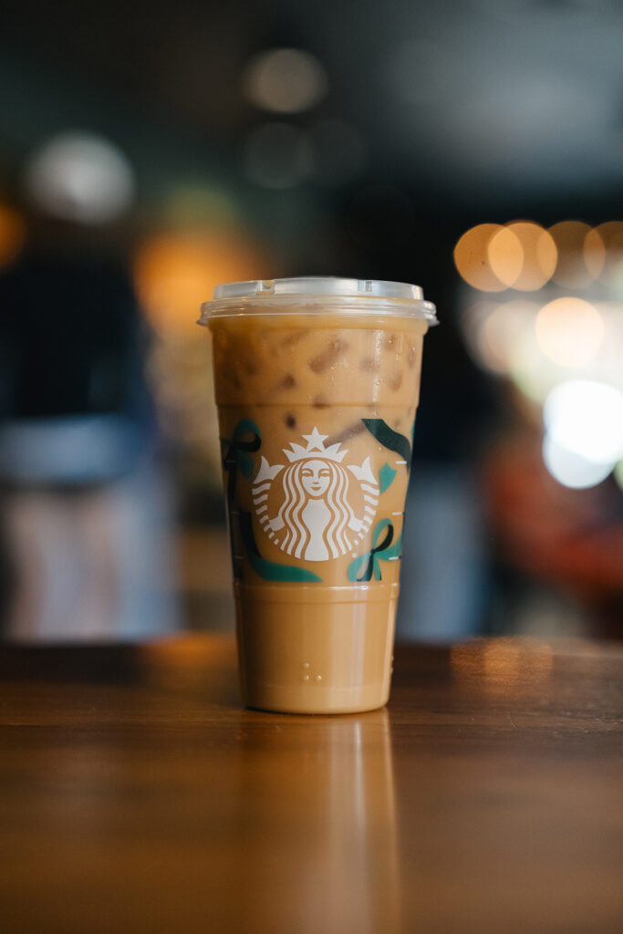 A clear cup filled with iced coffee sits on a wooden table, featuring a prominent logo and decorative elements. Blurred background lights create a cozy atmosphere.