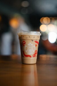 An iced coffee drink in a clear cup with a lid, featuring the Starbucks logo and decorative red ribbons, sits on a wooden table. The background is softly blurred with warm bokeh lights.