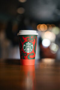 A festive Starbucks coffee cup with a red and green design featuring bows and the Starbucks logo, placed on a wooden surface.