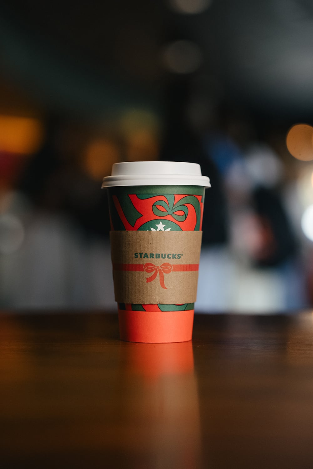 A festive Starbucks coffee cup with a red and green design, featuring a paper sleeve, sits on a wooden table.