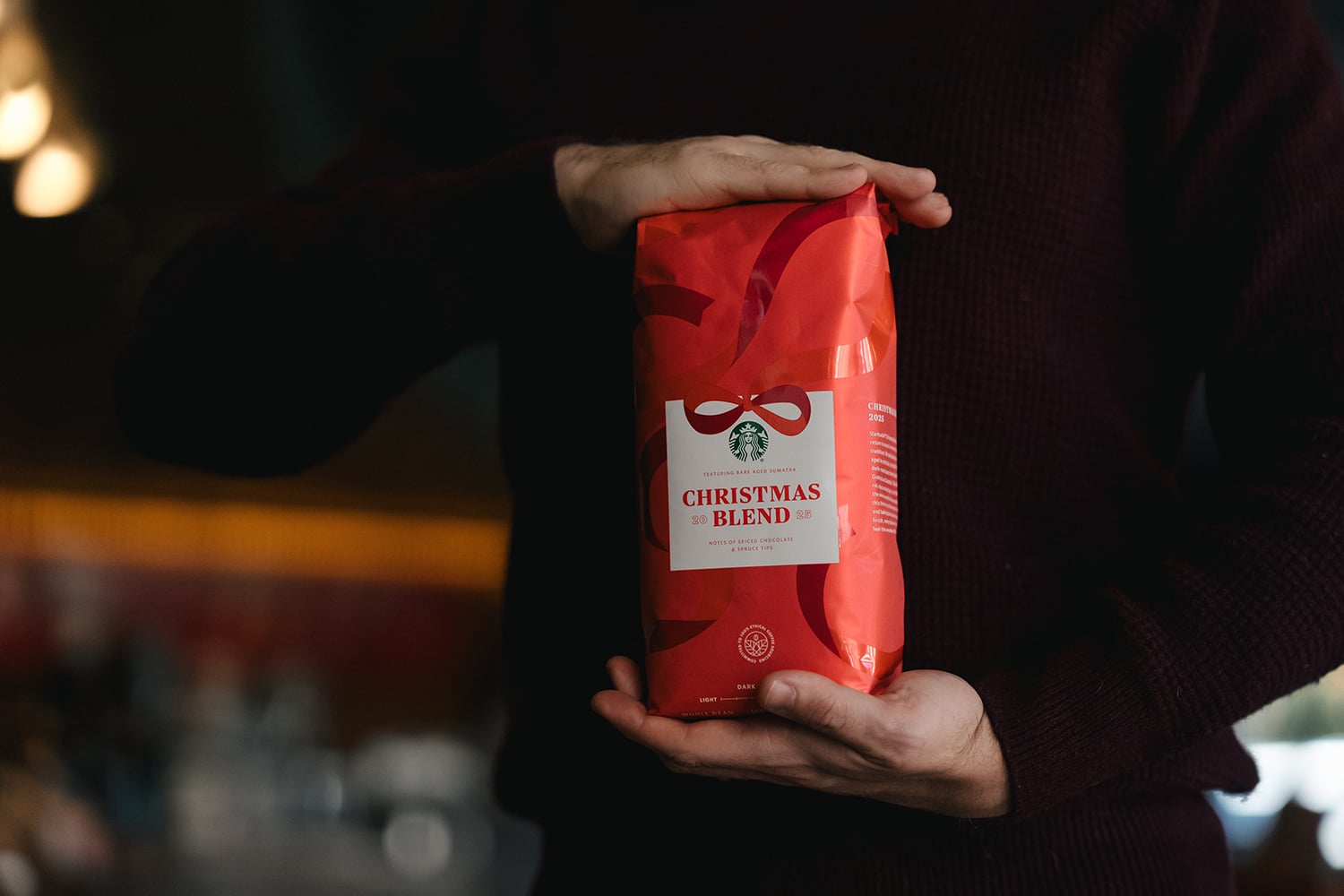 A person holds a red bag of Starbucks Christmas Blend coffee. The bag features a festive design with a bow and includes tasting notes.