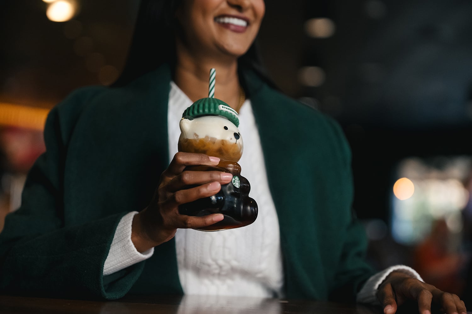 A person holds a bear-shaped cup with a green hat, filled with a beverage. They wear a green coat and a white sweater, smiling.