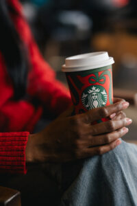 A person holds a festive Starbucks cup with a red and green design, wearing a red sweater and sitting casually.
