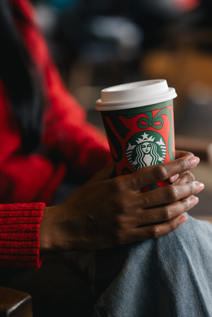 A person holds a festive Starbucks cup with a red and green design, wearing a red sweater and sitting casually.
