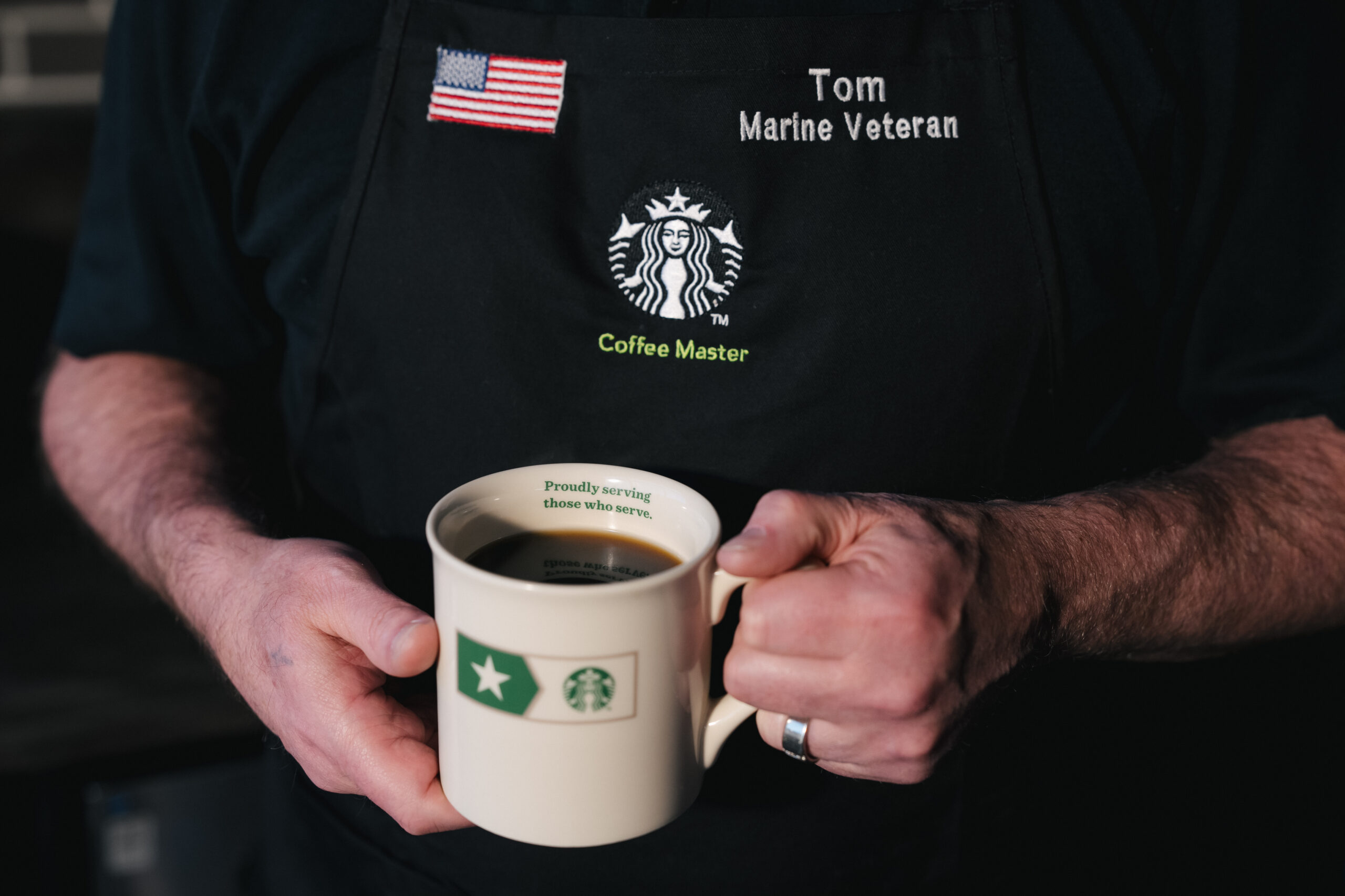 A person holds a coffee mug with a green star and Starbucks logo. They wear a black apron with "Coffee Master" and "Marine Veteran" embroidered, along with an American flag. The mug's interior reads, "Proudly serving those who serve.