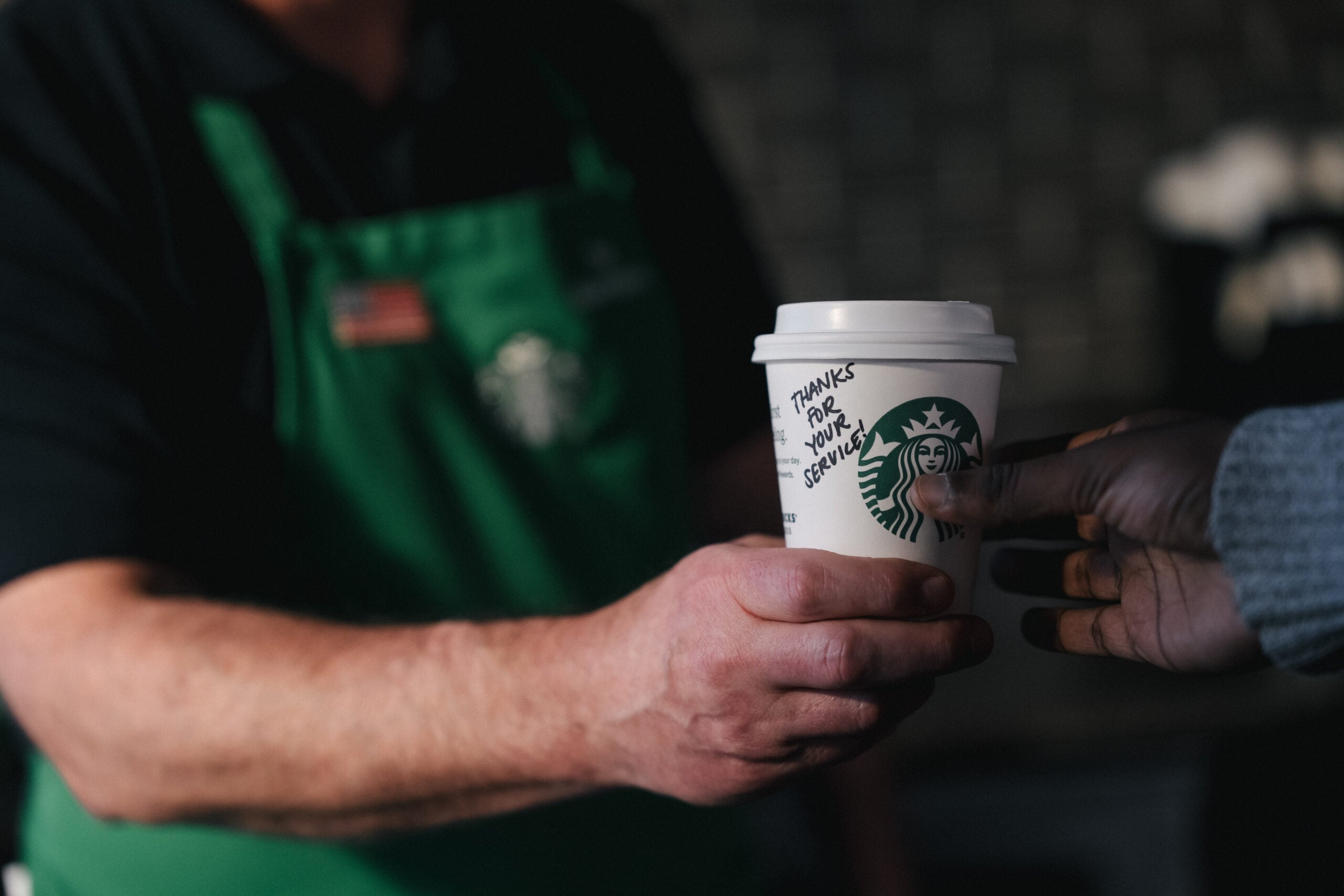 A barista in a green apron hands a coffee cup to a customer. The cup has a message written on it: "THANKS FOR YOUR SERVICE!