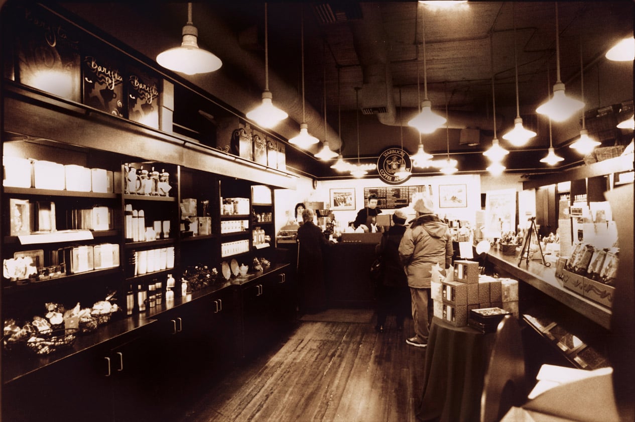 A vintage-style coffee shop interior with wooden shelves displaying various products. Customers are at the counter, and warm lighting creates a cozy atmosphere.