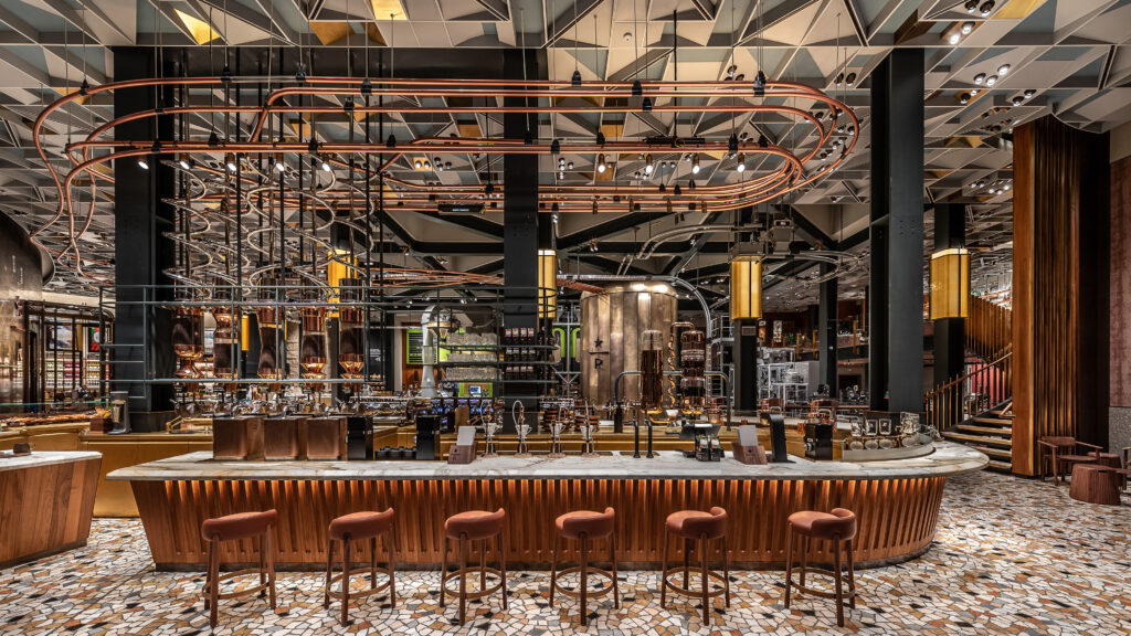 A modern bar interior featuring a long, curved counter with brown stools. Copper pipes and industrial elements are prominent, with a large brewing apparatus in the background. The ceiling has a geometric design, and the floor is decorated with mosaic tiles.