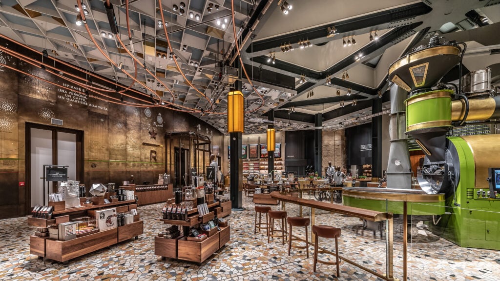 A modern café interior featuring wooden display shelves, a mosaic floor, and stylish lighting. The space is well-lit with a contemporary design.