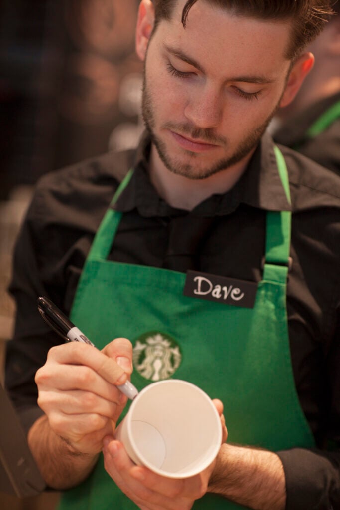 A barista in a green apron is focused on writing on a white cup with a black marker. The background is blurred, emphasizing the action.