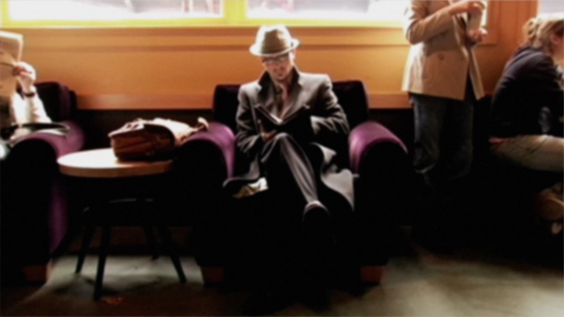 A man in a stylish coat and hat sits in a purple armchair, reading a book. Nearby, a woman stands holding a drink.