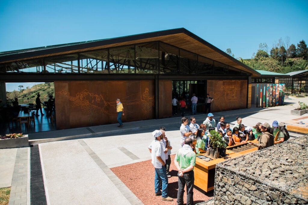 A modern building with a sloped roof features a large, rust-colored wall with etched designs. A group of people gathers around a wooden table displaying plants, while others are seen entering the building. The scene is set under a clear blue sky.