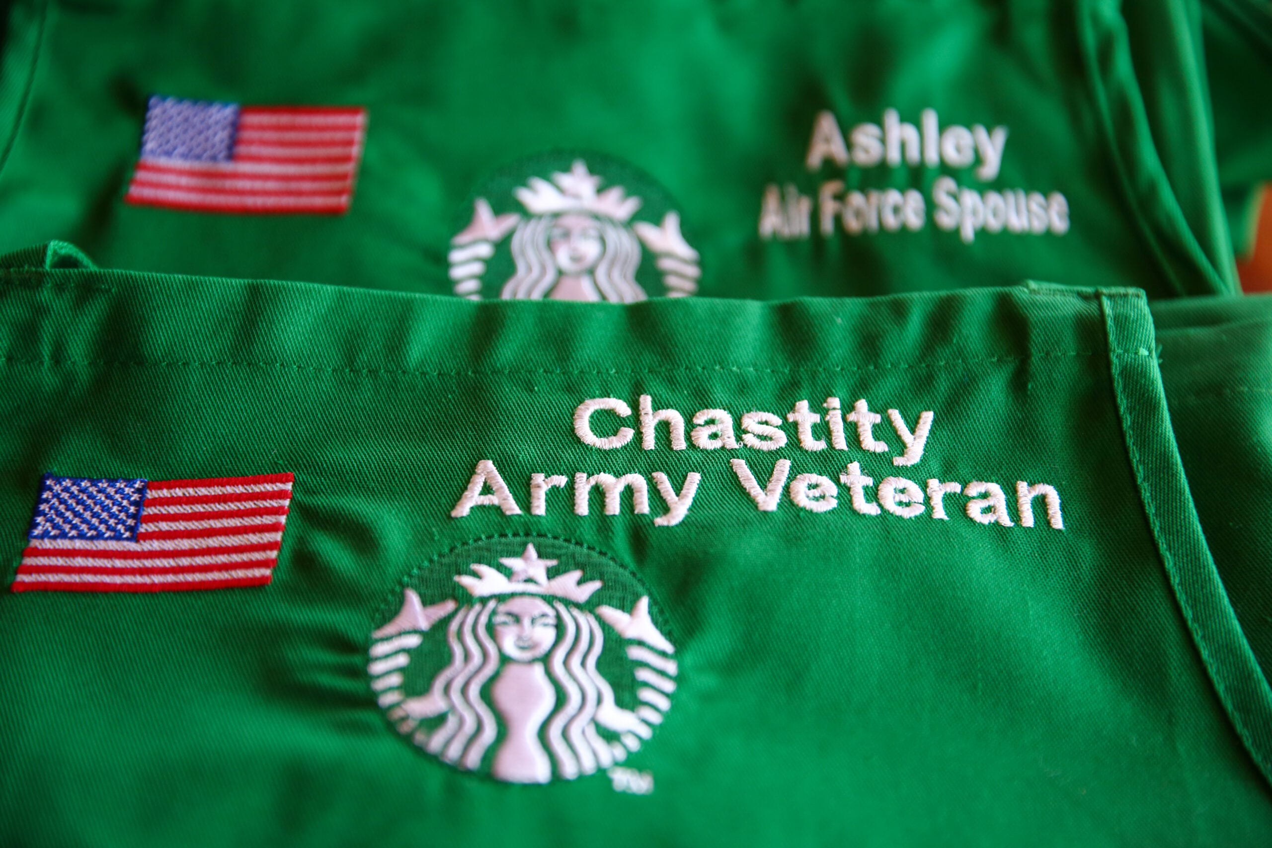 Two green aprons are displayed, featuring embroidered names and titles. One reads "Ashley, Air Force Spouse," and the other "Chastity, Army Veteran," alongside the Starbucks logo and a small American flag.