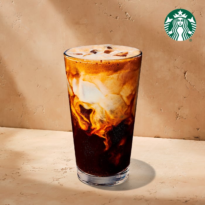 A tall glass filled with iced coffee, featuring swirls of cream and dark liquid, set against a textured beige background.