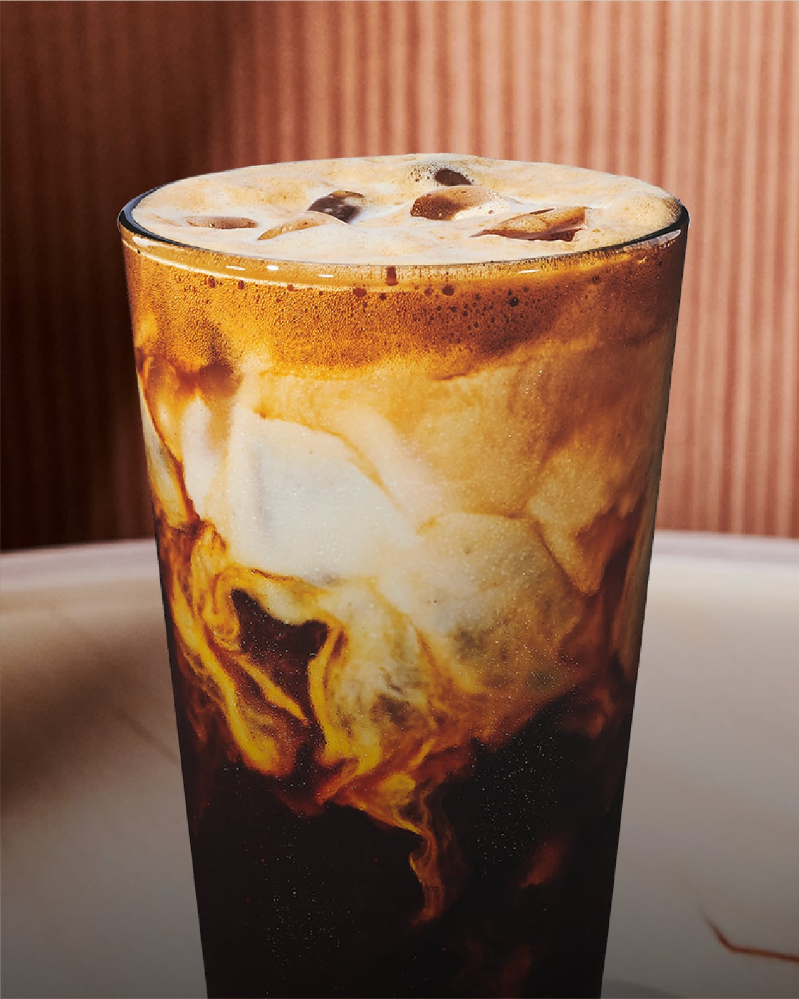 A glass filled with iced coffee, featuring a creamy swirl of milk blending with dark coffee. The background has a warm, textured tone.
