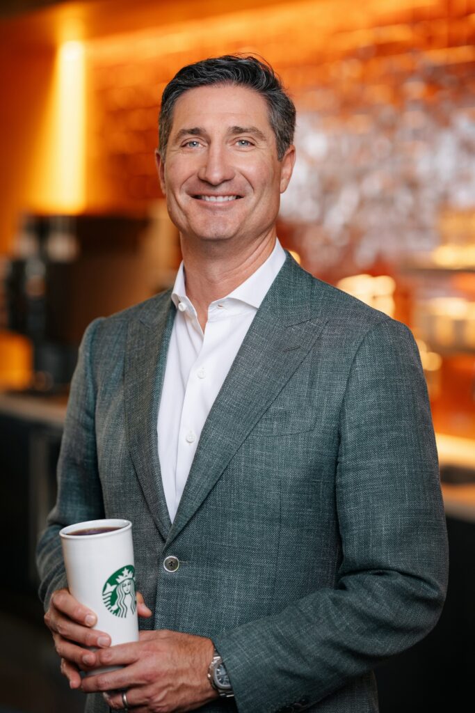 A man in a gray blazer holds a Starbucks cup, smiling confidently. The background features warm lighting and a coffee shop setting.