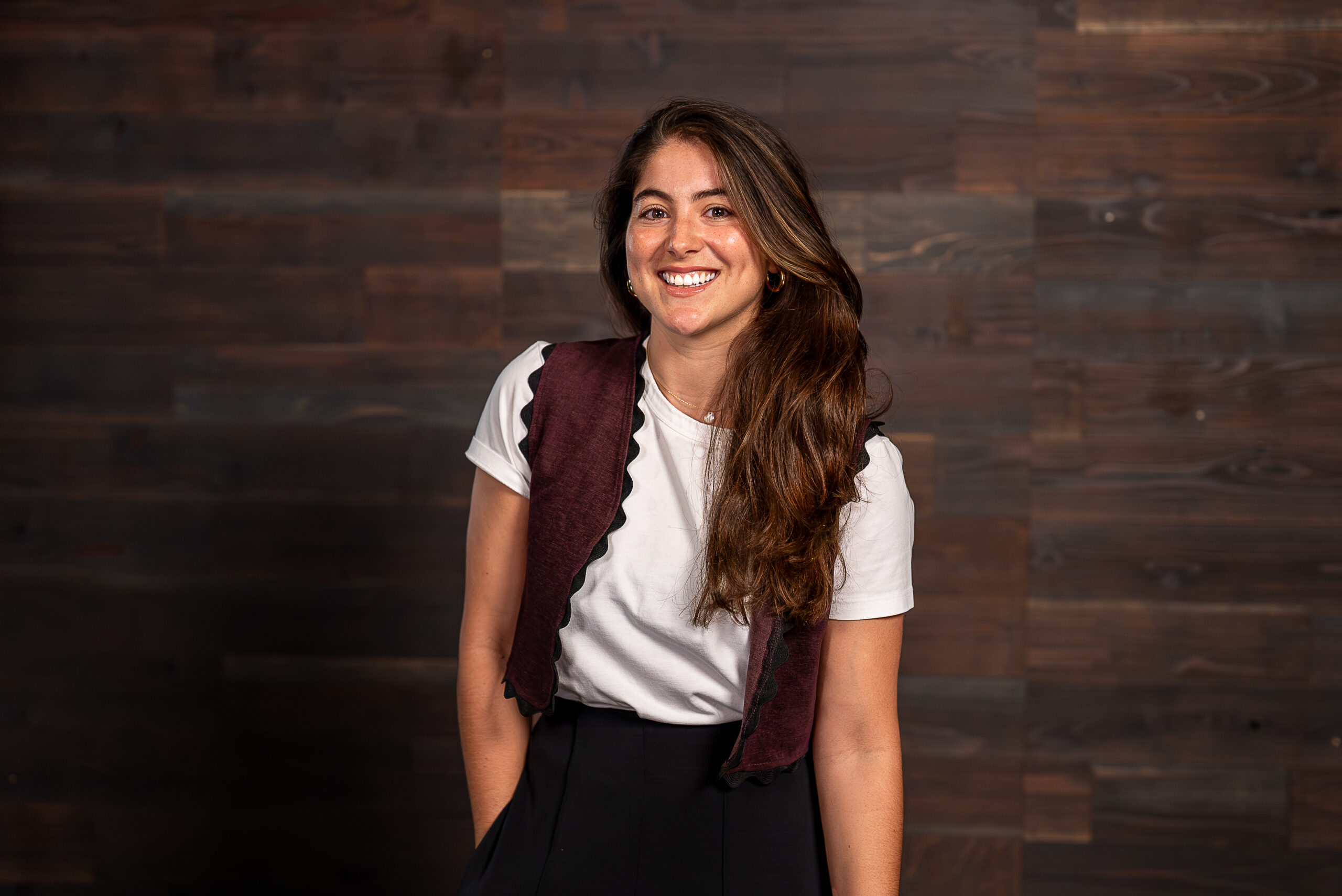 A woman with long, wavy hair smiles while wearing a white t-shirt and a burgundy vest with scalloped edges. The background features wooden panels.
